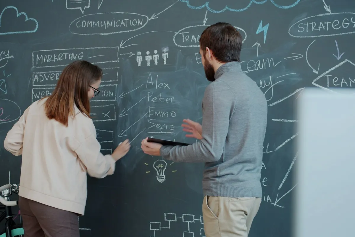 Two colleagues map a workflow together on a chalkboard